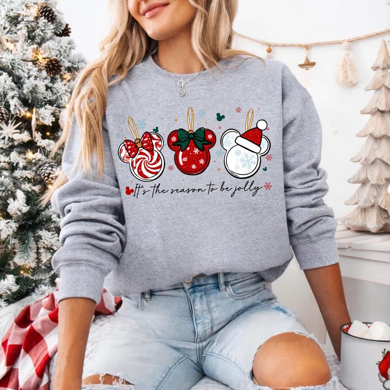 May include: A gray sweatshirt featuring three Mickey Mouse-shaped Christmas ornaments. The ornaments are red, white, and green, with a candy cane, bow, and Santa hat design. The text "It's the season to be jolly" is printed below the ornaments.