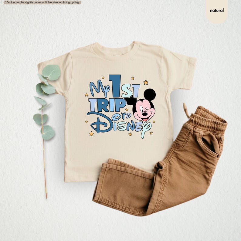 K&ouml;nnte beinhalten: Ein beiges T-Shirt mit einer blauen und wei&szlig;en Grafik, auf der "My 1st Trip to Disney" steht, sowie einem Cartoon-Bild von Mickey Mouse. Das Shirt liegt auf einem wei&szlig;en Hintergrund mit einer braunen Hose und einer gr&uuml;nen Pflanze.