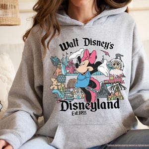 May include: Sport grey hoodie featuring Minnie Mouse with the text "Walt Disney's Disneyland Est.1955". The design includes various Disneyland attractions. The hoodie has a front pocket and a drawstring hood.