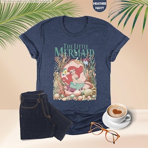 May include: Heather navy t-shirt featuring a colorful graphic of Ariel from The Little Mermaid. The design includes the text "THE LITTLE MERMAID" above the illustration. A pair of dark wash jeans and a cup of coffee with heart-shaped foam are also in the image.