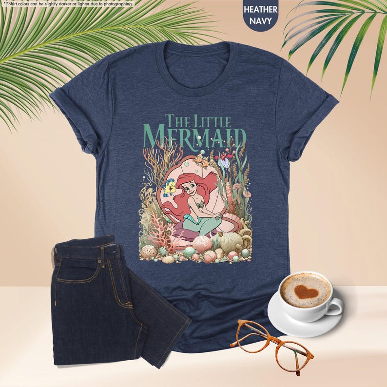 May include: Heather navy t-shirt featuring a colorful graphic of Ariel from The Little Mermaid. The design includes the text "THE LITTLE MERMAID" above the illustration. A pair of dark wash jeans and a cup of coffee with heart-shaped foam are also in the image.