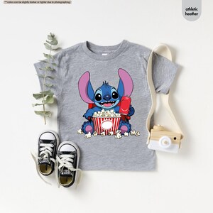 May include: Athletic heather gray t-shirt featuring a cartoon character holding popcorn and a drink. The character is blue with large ears and is sitting in front of a red and white striped popcorn bucket. Black and white sneakers are next to the shirt.