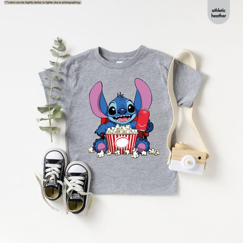 May include: Athletic heather gray t-shirt featuring a cartoon character holding popcorn and a drink. The character is blue with large ears and is sitting in front of a red and white striped popcorn bucket. Black and white sneakers are next to the shirt.