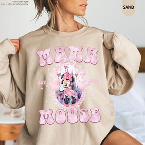 May include: Sand-colored sweatshirt with the words "MAMA MOUSE" in pink bubble letters. A pink framed graphic features Minnie Mouse in a pink dress. The text "EST 2020" is also visible. The sweatshirt is a casual style.