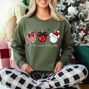 May include: Military green sweatshirt with three Mickey Mouse-shaped Christmas ornaments. The ornaments are red, white, and green, with candy cane, bow, and Santa hat designs. The text "It's the season to be jolly" is below the ornaments.