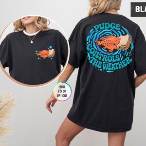 Puede incluir: Camiseta negra con un gr&aacute;fico colorido de un pez naranja con un s&aacute;ndwich, rodeado de olas azules y el texto "Pudge Controls The Weather" en la espalda. Una versi&oacute;n m&aacute;s peque&ntilde;a del gr&aacute;fico est&aacute; en la parte delantera.