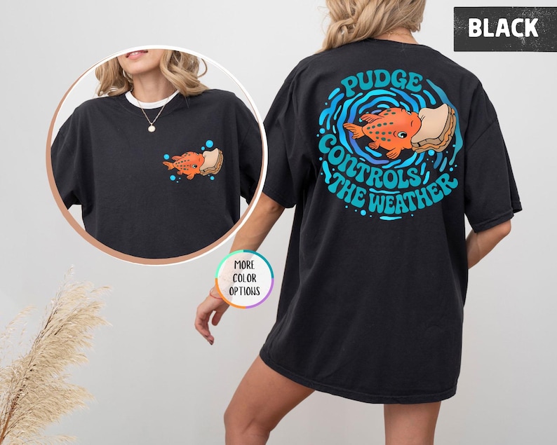 Puede incluir: Camiseta negra con un gr&aacute;fico colorido de un pez naranja con un s&aacute;ndwich, rodeado de olas azules y el texto "Pudge Controls The Weather" en la espalda. Una versi&oacute;n m&aacute;s peque&ntilde;a del gr&aacute;fico est&aacute; en la parte delantera.