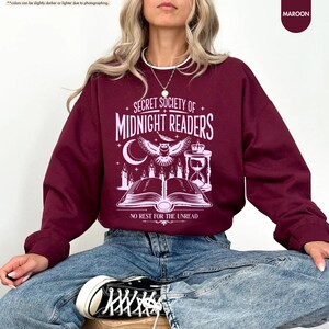 May include: Maroon sweatshirt with the text "SECRET SOCIETY OF MIDNIGHT READERS" and an owl graphic. The design includes a crescent moon, candles, an hourglass, and an open book. The phrase "NO REST FOR THE UNREAD" is below the book.