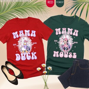 May include: Two t-shirts, one red and one forest green, with the text "MAMA DUCK" and "MAMA MOUSE" in a retro font. Each shirt features a cartoon character illustration. A pair of black flats and jeans are also visible.