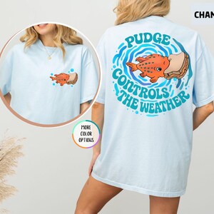 Puede incluir: Camiseta azul claro con un dise&ntilde;o de pez de dibujos animados. La parte delantera de la camiseta presenta un peque&ntilde;o gr&aacute;fico de pez y s&aacute;ndwich. La parte trasera de la camiseta tiene un dise&ntilde;o m&aacute;s grande con el texto "Pudge Controls The Weather" en verde azulado.