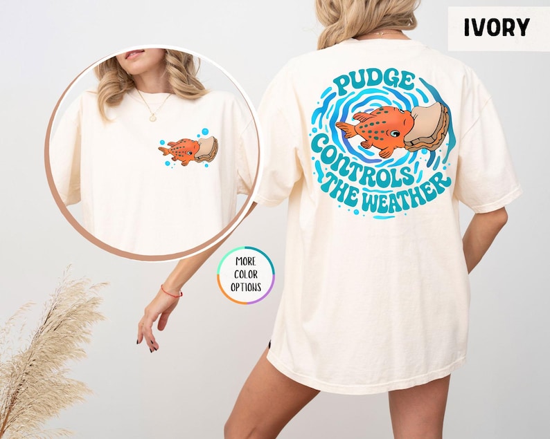Puede incluir: Camiseta de color marfil con un dise&ntilde;o de pez de dibujos animados. La parte delantera de la camiseta tiene un peque&ntilde;o gr&aacute;fico de pez, mientras que la parte trasera muestra un dise&ntilde;o m&aacute;s grande con el texto "Pudge Controls the Weather" en azul. La camiseta es de corte holgado.