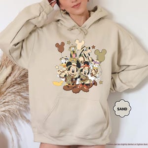 May include: Sand-colored hoodie featuring a graphic of Disney characters, including Mickey and Minnie Mouse, Donald Duck, and Goofy, in safari attire. The design includes Mickey Mouse-shaped balloons and stars. The hoodie has a kangaroo pocket and a drawstring hood.