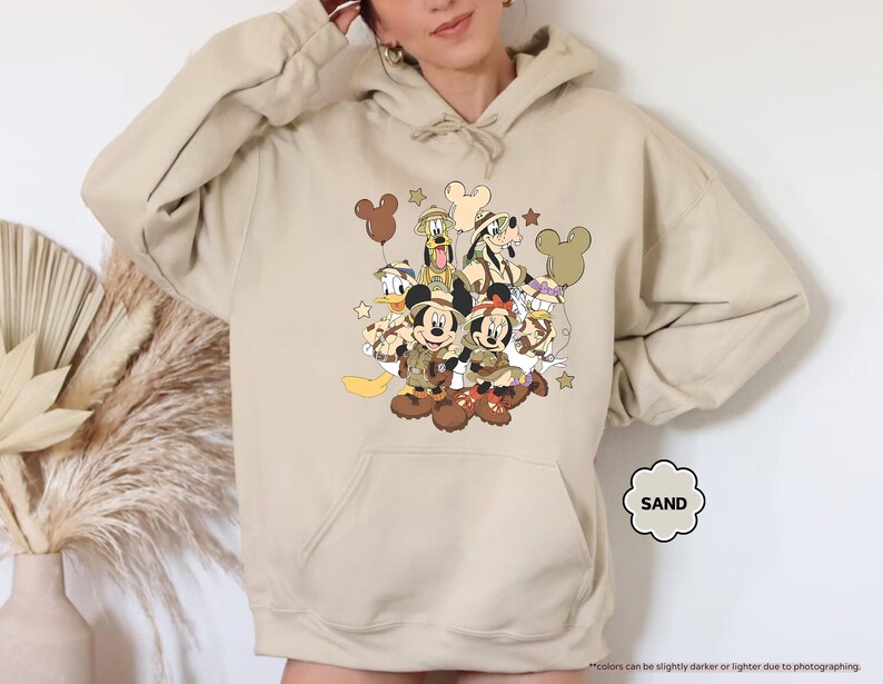 May include: Sand-colored hoodie featuring a graphic of Disney characters, including Mickey and Minnie Mouse, Donald Duck, and Goofy, in safari attire. The design includes Mickey Mouse-shaped balloons and stars. The hoodie has a kangaroo pocket and a drawstring hood.