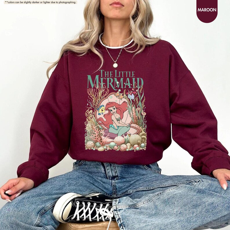 May include: Maroon sweatshirt featuring a graphic of Ariel from The Little Mermaid. The design includes the text "THE LITTLE MERMAID" in a stylized font, with Ariel surrounded by underwater elements. The sweatshirt is a solid maroon color.