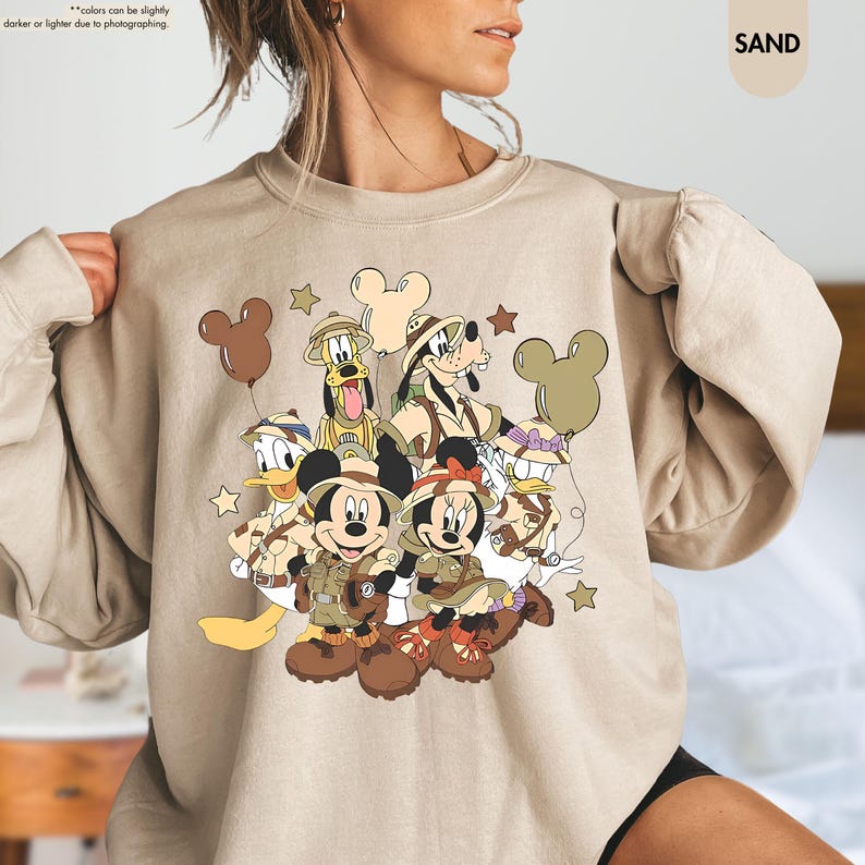 May include: Sand-colored sweatshirt featuring a graphic of Mickey Mouse, Minnie Mouse, Donald Duck, Goofy, and Pluto in safari outfits. The design includes brown Mickey Mouse-shaped balloons and stars. The word "SAND" is printed on the upper right.