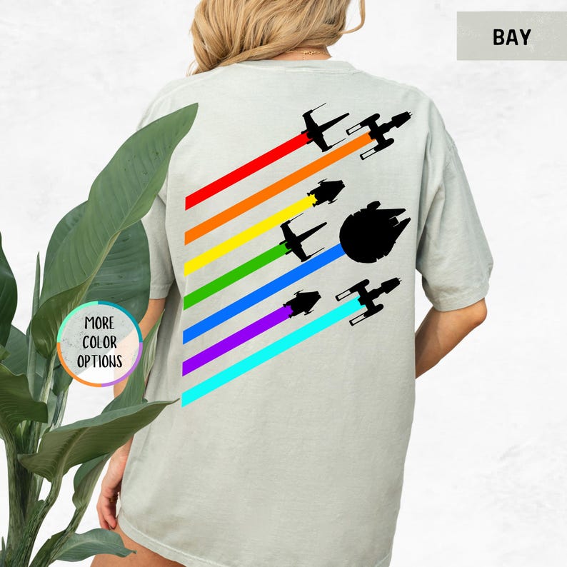 K&ouml;nnte beinhalten: Ein hellgraues T-Shirt mit einem Regenbogenmuster und Silhouetten von Star Wars Raumschiffen. Der Text "BAY" befindet sich in der oberen rechten Ecke des Bildes.