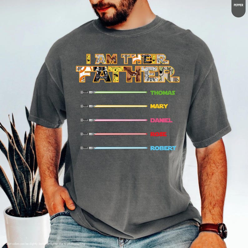 Puede incluir: Camiseta gris oscuro con el texto "I AM THE FATHER" en un dise&ntilde;o de patchwork. Debajo, cinco sables de luz de colores con los nombres: Thomas, Mary, Daniel, Rose y Robert. La camiseta es de estilo casual.