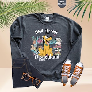 May include: Dark heather gray sweatshirt featuring a graphic of Pluto from Walt Disney's Disneyland. The design includes the text "Walt Disney's Disneyland Est. 1955" and various park icons. The sweatshirt is paired with dark blue jeans and orange sneakers.