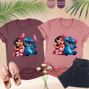 K&ouml;nnte beinhalten: Zwei Heather-T-Shirts, eines in Mauve und eines in Maroon, mit einem Cartoon-Bild von Lilo und Stitch. Das Bild zeigt Lilo und Stitch zusammen sitzend. Ein Paar rosa Turnschuhe und eine dunkelblaue Jeans sind ebenfalls im Bild.