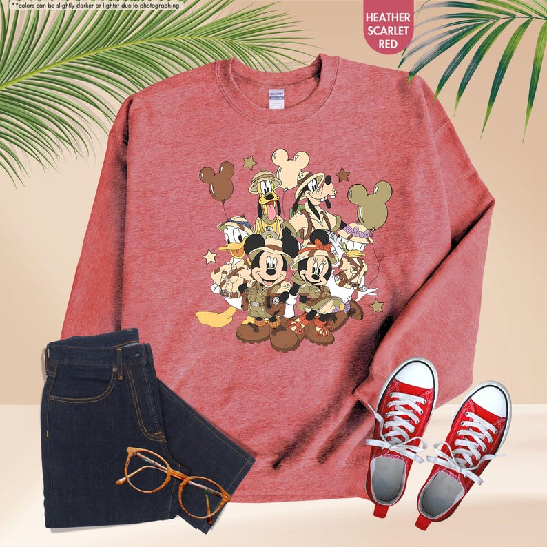 May include: A heather scarlet red sweatshirt featuring a cartoon graphic of Mickey Mouse and friends in safari outfits. The image includes a pair of dark blue jeans, red sneakers, and gold-rimmed glasses. The text "HEATHER SCARLET RED" is visible.