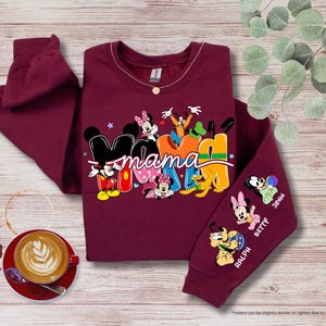 K&ouml;nnte beinhalten: Burgunderfarbenes Sweatshirt mit dem Wort "Mama" in bunten Buchstaben, mit Micky Maus und Freunden. Der &Auml;rmel zeigt Zeichentrickfiguren und Namen. Eine Kaffeetasse und dekorative Elemente befinden sich im Hintergrund.