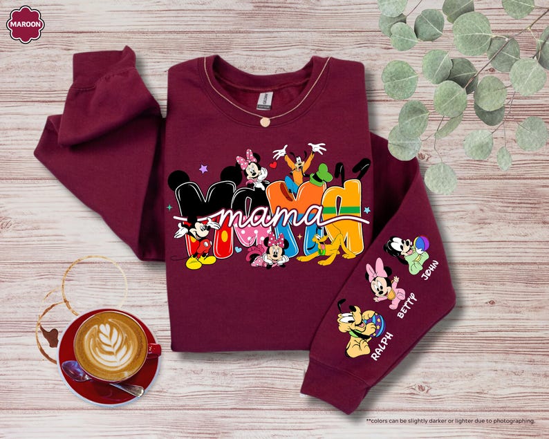 K&ouml;nnte beinhalten: Burgunderfarbenes Sweatshirt mit dem Wort "Mama" in bunten Buchstaben, mit Micky Maus und Freunden. Der &Auml;rmel zeigt Zeichentrickfiguren und Namen. Eine Kaffeetasse und dekorative Elemente befinden sich im Hintergrund.