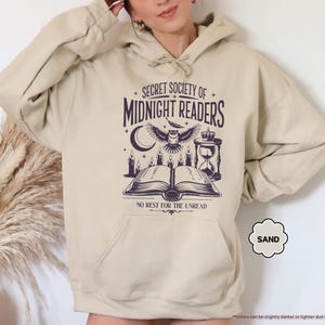 May include: Sand-colored hoodie with a front graphic. The graphic features the text "SECRET SOCIETY OF MIDNIGHT READERS" above an owl, open book, candles, and hourglass. The text "NO REST FOR THE UNREAD" is below the book.