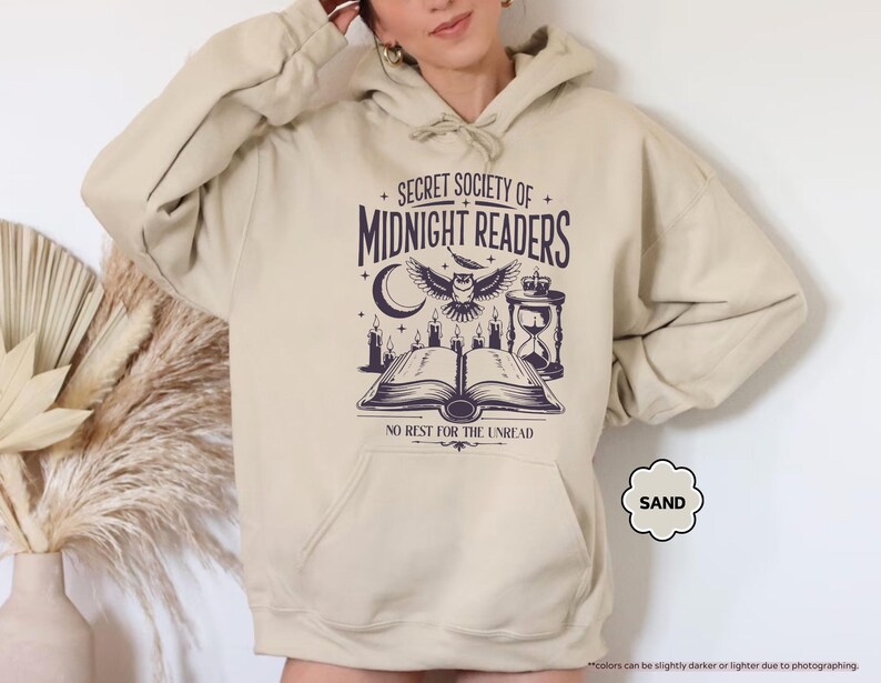 May include: Sand-colored hoodie with a front graphic. The graphic features the text "SECRET SOCIETY OF MIDNIGHT READERS" above an owl, open book, candles, and hourglass. The text "NO REST FOR THE UNREAD" is below the book.