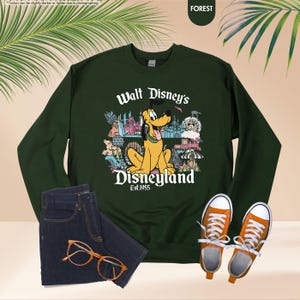 May include: A dark green sweatshirt featuring Pluto from Disney's Disneyland, with the text "Walt Disney's Disneyland Est. 1955". The image also shows a pair of blue jeans, orange-colored glasses, and orange sneakers.