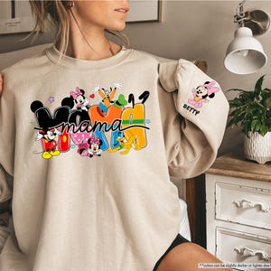 K&ouml;nnte beinhalten: Beiges Sweatshirt mit dem Wort "Mama" in bunten Blockbuchstaben, mit den Disney-Figuren Mickey Mouse, Minnie Mouse, Goofy und Pluto. Der &Auml;rmel hat eine Minnie Mouse Grafik und den Namen "Betty".