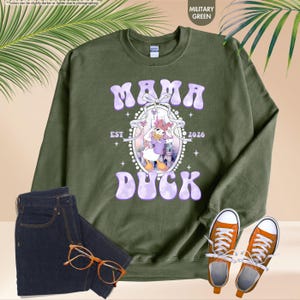 May include: Military green sweatshirt with the text "MAMA DUCK EST 2026" in a purple, retro font. The design includes a cartoon duck character. The image also shows a pair of dark blue jeans, orange sneakers, and eyeglasses.