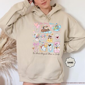 May include: Sand-colored hoodie featuring a colorful graphic of Disney characters and icons. The design includes Mickey and Minnie Mouse, Dumbo, and other elements. Text on the hoodie reads "The Most Magical Place on Earth."