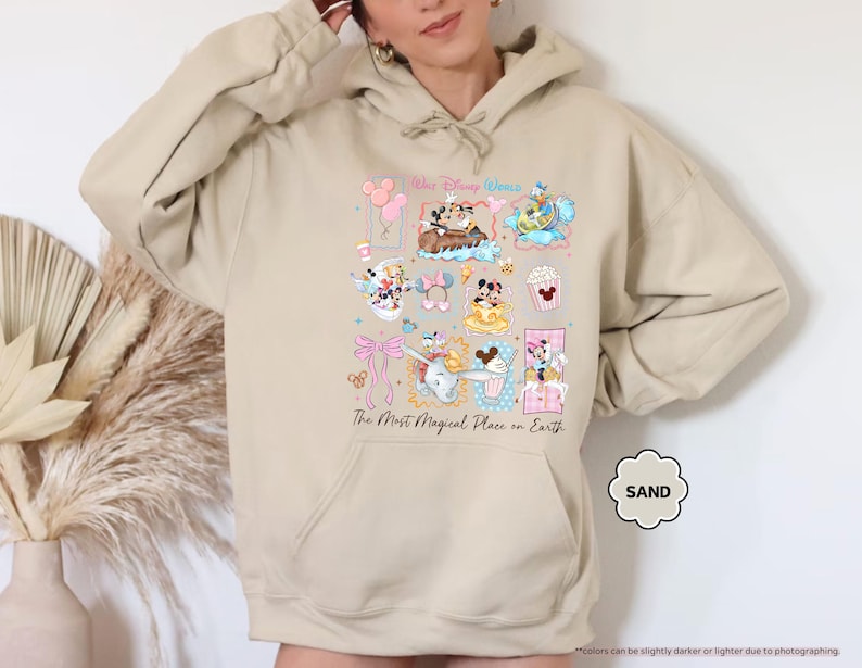 May include: Sand-colored hoodie featuring a colorful graphic of Disney characters and icons. The design includes Mickey and Minnie Mouse, Dumbo, and other elements. Text on the hoodie reads "The Most Magical Place on Earth."