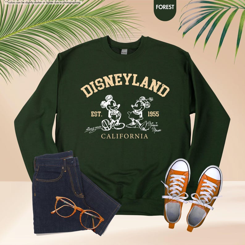 May include: A dark green sweatshirt featuring the word "DISNEYLAND" and an image of Mickey and Minnie Mouse. The text "EST. 1955 CALIFORNIA" is below the image. The sweatshirt is styled with blue jeans, orange trainers, and glasses.