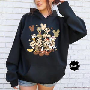 May include: Black hooded sweatshirt featuring a graphic of Disney characters, including Mickey and Minnie Mouse, Donald Duck, and Goofy, in safari attire. The design includes stars and Mickey Mouse silhouettes. The word "BLACK" is in a black circle.