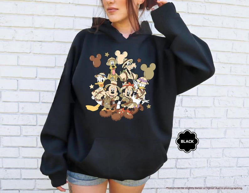 May include: Black hooded sweatshirt featuring a graphic of Disney characters, including Mickey and Minnie Mouse, Donald Duck, and Goofy, in safari attire. The design includes stars and Mickey Mouse silhouettes. The word "BLACK" is in a black circle.