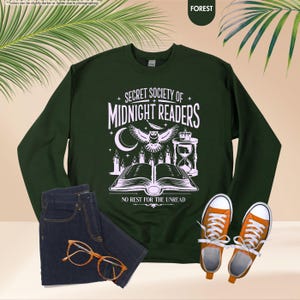 May include: A forest green sweatshirt with the text "SECRET SOCIETY OF MIDNIGHT READERS" and an illustration of an owl, open book, candles, and hourglass. The sweatshirt is paired with dark blue jeans and orange sneakers.