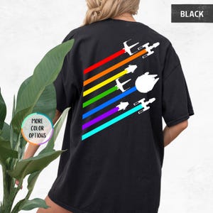 K&ouml;nnte beinhalten: Schwarzes T-Shirt mit einem Regenbogenstreifen-Design und wei&szlig;en Silhouetten von Star Wars-Raumschiffen, die &uuml;ber die Streifen fliegen. Der Text "BLACK" befindet sich in der oberen rechten Ecke des Bildes.