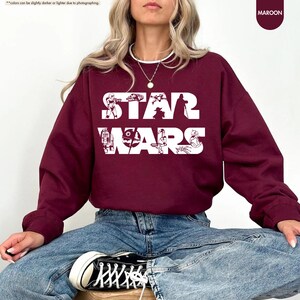 May include: Maroon sweatshirt with the words "STAR WARS" in white block letters. The sweatshirt features white illustrations of characters and elements from the Star Wars franchise. The model is wearing blue jeans and black sneakers.