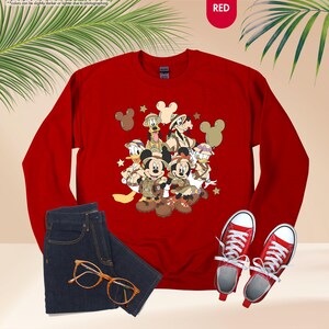 May include: A red crewneck sweatshirt featuring a graphic of Disney characters in safari outfits. The image includes a pair of dark wash jeans, red sneakers, and tortoiseshell glasses. The word "RED" is in the upper right corner.