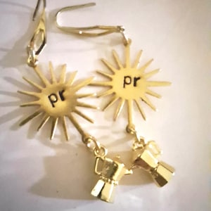 May include: Gold-colored earrings featuring sunburst charms with the letters "pr" and miniature coffee pot charms. The earrings have a hook closure and are designed to dangle.
