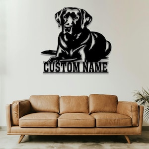 May include: Black metal silhouette of a Labrador Retriever dog lying down with the text "CUSTOM NAME" below. The silhouette is mounted on a white wall above a brown leather couch.