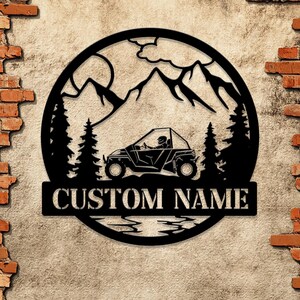 Könnte beinhalten: Schwarze Metallsilhouette einer Bergszene mit einer Person, die ein Side-by-Side-Fahrzeug fährt. Die Silhouette befindet sich in einem Kreis mit einem See unten und dem Text "CUSTOM NAME" unten.