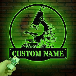 May include: A black metal silhouette of a microscope with floral details, illuminated in green. The text "CUSTOM NAME" is displayed on a black banner below the microscope.
