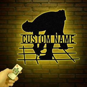 May include: A black silhouette of a person laying down concrete with a rebar grid underneath. The silhouette is illuminated with a yellow light. The text "CUSTOM NAME" is above the silhouette. A remote control is in the lower left corner of the image.