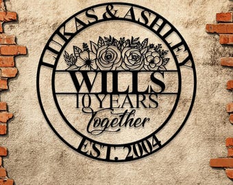 Könnte beinhalten: Ein rundes Wand-Schild aus schwarzem Metall mit dem Text LUKAS & ASHLEY oben, einem Blumenmotiv in der Mitte und dem Text WILLS, 10 YEARS Together sowie EST. 2004 darunter. Das Schild hat ein ausgeschnittenes Design mit klarer Schrift.