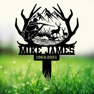 May include: Black metal silhouette of a hunter with a rifle, two deer, and a mountain scene. The silhouette is mounted on a metal stake with the name "Mike James" and the dates "1963-2024" below.