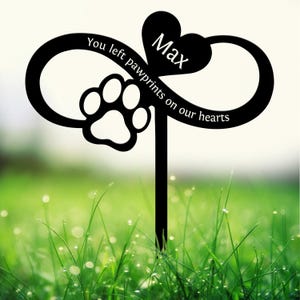 May include: Black metal garden stake featuring an infinity symbol, a paw print, and a heart with the name "Max." The text reads "You left pawprints on our hearts." The stake is set in a green grassy area.