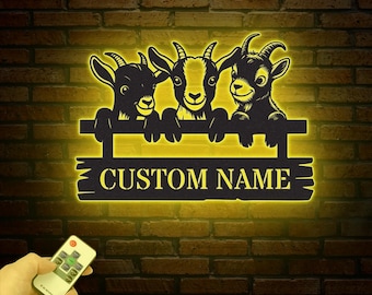 May include: A black metal silhouette of three goats peeking over a fence with a custom name sign. The sign is illuminated with a warm yellow light.