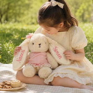 May include: A cream-colored plush rabbit toy featuring long ears with embroidered names Naneri and Simon in pink thread. The rabbit wears a light pink dress with a ruffled hem and a small pink flower accessory attached to the top of its head.